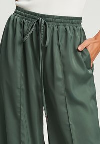 Groene satijnen broek met wijde pijpen, een tailleband met trekkoord en zijzakken. Soepele textuur en relaxte pasvorm, met een subtiele glans.