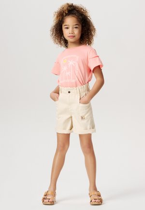 Jeune fille aux cheveux bouclés portant un T-shirt rose avec des palmiers, un short beige taille haute et des sandales beiges, se tenant les mains dans les poches.