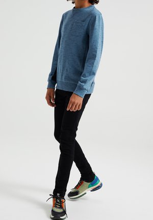 Sudadera de cuello redondo textura azul claro con un bolsillo en el pecho, combinada con vaqueros negros de corte ajustado y zapatillas deportivas multicolor.