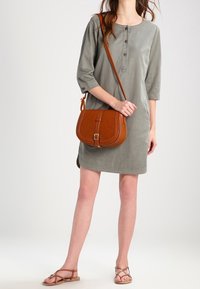 Nainen, joka pukeutuu harmaaseen kolmen neljäsosapituiseen mekkoon, mukana ruskea olkalaukku ja ruskeat remmisandaalit, seisoo yksinkertaista taustaa vasten.