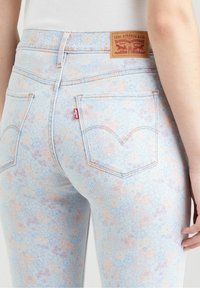 Pantalones vaqueros de estampado floral en azul claro con un acabado de denim texturizado, que presentan una etiqueta de cuero marrón y un logotipo rojo de la marca en el bolsillo trasero.