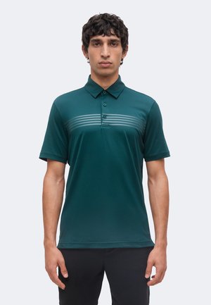 Homme portant un polo vert à manches courtes avec des rayures horizontales sur la poitrine, debout devant un fond clair et uni.