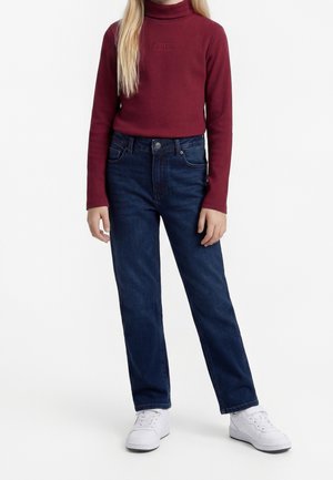 Person iført en rød rullekravesweater, mørkeblå jeans og hvide sneakers, stående mod en ensfarvet hvid baggrund.