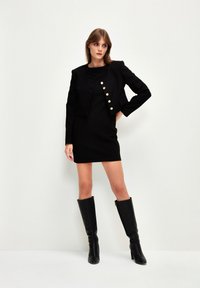 Černé upnuté šaty v kombinaci s krátkým černým sakem s zlatými knoflíky. Modelka má na sobě černé kozačky do kolen; hladké textury.