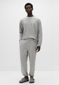 Szary długi top z długim rękawem oraz pasujące spodnie dresowe z elastycznymi mankietami, wykonane z miękkiego materiału. Model nosi szare wsuwane buty z paskiem.