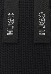 Tessuto a maglia nero con due etichette tessute nere che mostrano il marchio "HUGO" in testo bianco, caratterizzate da un design testurizzato.