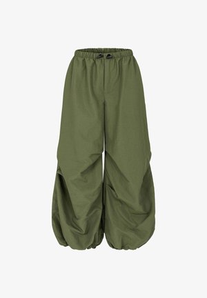 Kobieta o kręconych włosach stoi uśmiechnięta na zewnątrz, ubrana w czarny t-shirt z grafiką, luźne oliwkowe spodnie i sandały, za nią rośliny i palma.