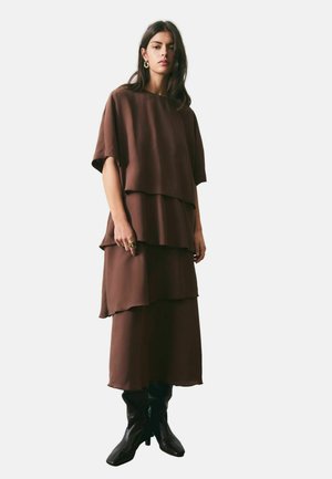 Kvinde iført en lagdelt brun kjole med korte ærmer og sorte støvler til midt på læggen, stående mod en ensfarvet lys baggrund.