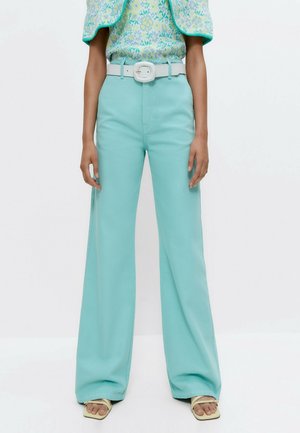 Femme portant un pantalon large taille haute turquoise, une ceinture blanche, un haut à fleurs et des sandales beiges à bout ouvert, debout devant un fond blanc.