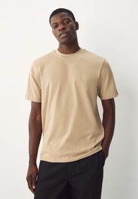 T-shirt beige in cotone con scollo rotondo e maniche corte, caratterizzata da una vestibilità rilassata e una texture liscia su uno sfondo semplice.