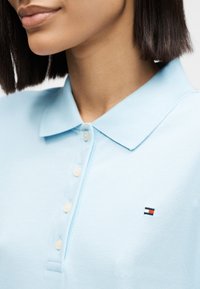 Κοντινό πλάνο ενός ατόμου που φορά ένα ανοιχτό μπλε πόλο με γιακά, τέσσερα κουμπιά και ένα μικρό κόκκινο, λευκό και ναυτικό λογότυπο στο στήθος.