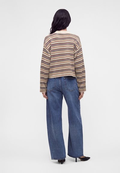 Femme portant un t-shirt rayé à manches longues, un jean bleu à jambes larges et des chaussures à talons noires, vue de dos sur un fond uni.