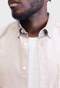 Jasnobeżowa koszula sztruksowa na guziki z klasycznym kołnierzykiem, z białymi guzikami i teksturowaną powierzchnią, noszona na prążkowanej białej podkoszulce.