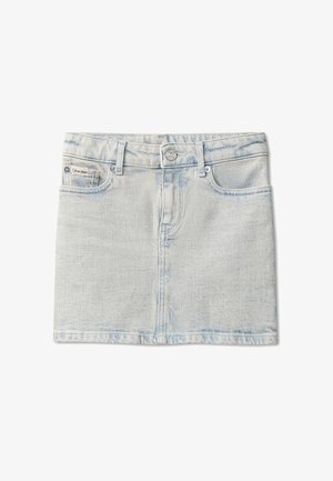 Jeune fille aux longs cheveux portant un sweat-shirt crème Calvin Klein, une jupe en denim clair, des chaussettes blanches et des baskets beige, debout les mains dans les poches.