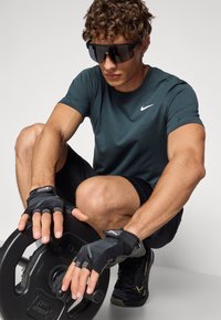 Musta painolaatta, jota henkilö pitää käsissään, yllään tummanvihreä Nike-paita ja mustat shortsit, sekä sormettomat hanskat ja tummat heijastavat aurinkolasit.