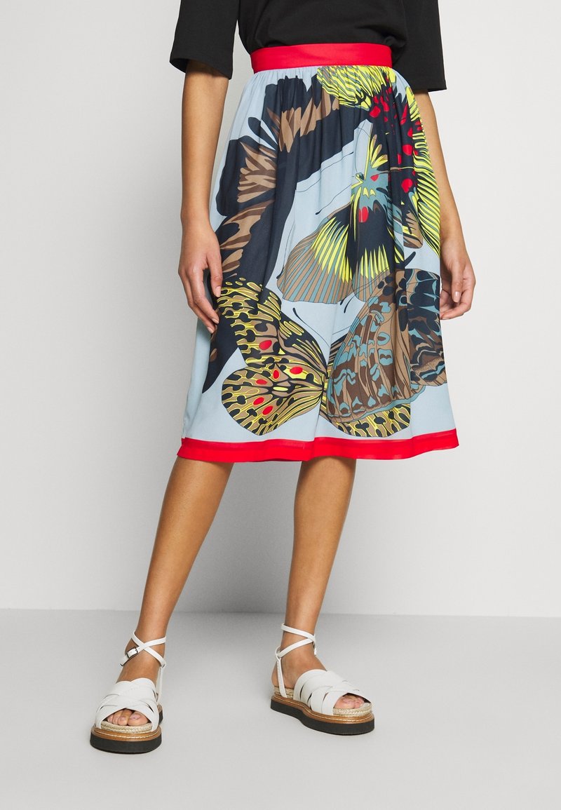Femme portant une jupe mi-longue avec un grand imprimé papillons multicolores, taille et ourlet rouges, associée à des sandales blanches à brides.