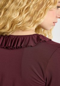 Bordeaux topp med ett texturerat tyg och volangdetalj vid halsringningen. Slät yta och figurnära design synlig på baksidan.