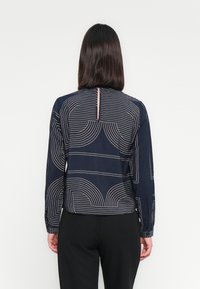 Marineblaue Bluse mit langen Ärmeln, die geometrische Linienmuster in Creme zeigt, mit einem Schlüsselrückendetail und leicht ausgestellt Ärmeln.