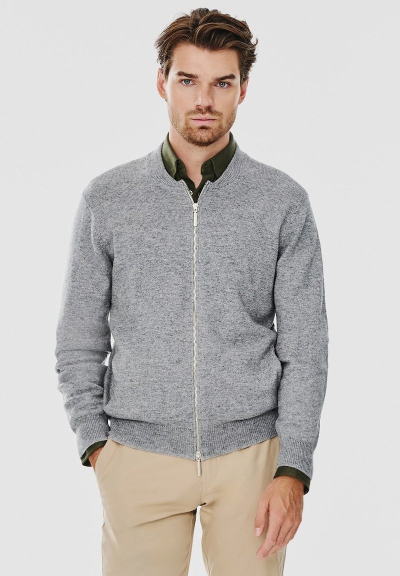 Szary sweter na zamek z okrągłym dekoltem i ściągaczami przy mankietach. Nosi się go na zielonej koszuli, zaprezentowany na jednolitym białym tle.