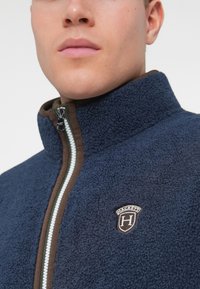 Námornícky fleecový kabát s vysokým golierom, hnedými zipsovými akcentmi a značkovou nášivkou. Podporuje textúrovanú tkaninu a kontrastné detaily zipsu.