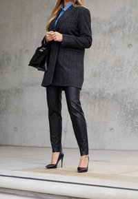 Kvinna klädd i en mörk rutig kavaj, blå skjorta, svarta läderbyxor och svarta högklackade skor, som håller i en svart handväska mot en betongvägg.