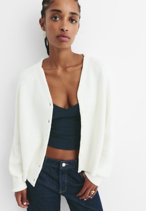 Weißer gerippter Cardigan mit übergroßen Ärmeln und Knopfverschluss, über einem dunkelblauen Oberteil getragen. Modell trägt dunkle Jeans.