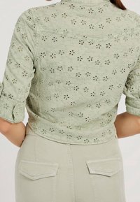 Light green embroidered floral lace shirt with rolled-up sleeves paired with high-waisted beige pants with flap back pockets.