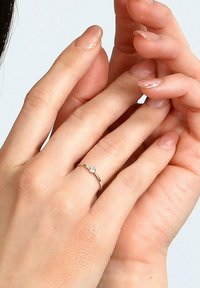 Goldring mit einem einzelnen runden Diamant-Akzent, ausgestattet mit einem schlanken Band. Die Hände sind zusammengelegt und zeigen einen natürlichen Nagel-Finish.