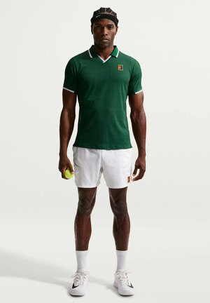 Mann trägt grünes Poloshirt, weiße Shorts und weiße Turnschuhe, hält einen Tennisball und steht vor einem schlichten hellen Hintergrund.