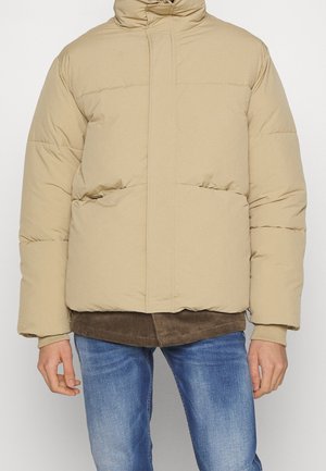 Giacca imbottita beige con colletto alto, cuciture orizzontali e due tasche frontali. Indossata sopra uno strato di velluto a coste marrone e jeans blu.