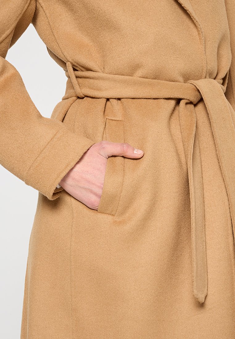 Beżowy płaszcz z mieszanki wełny z kołnierzem, paskiem i bocznymi kieszeniami. Miękka tekstura i dopasowany krój. Brak widocznych wzorów lub detali metalowych.