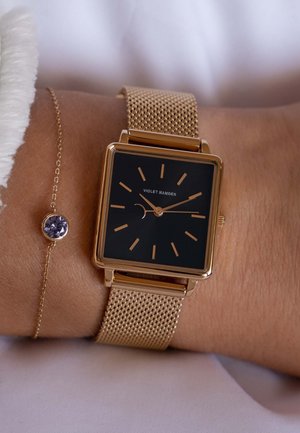 Montre bracelet en maille dorée avec un cadran carré noir, des index horaires dorés et des aiguilles. Accompagnée d'un délicat bracelet en chaîne dorée avec une pierre violette.