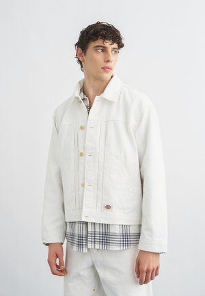 Młody mężczyzna z kręconymi włosami, ubrany w białą, zapinaną na guziki kurtkę na czarno-białą koszulę w kratę, na jednolitym białym tle.