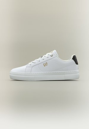 Zapatilla blanca de corte bajo con cordones blancos, logo dorado "GAP" en el lateral y lengüeta trasera negra con texto pequeño en naranja, sobre fondo neutro.