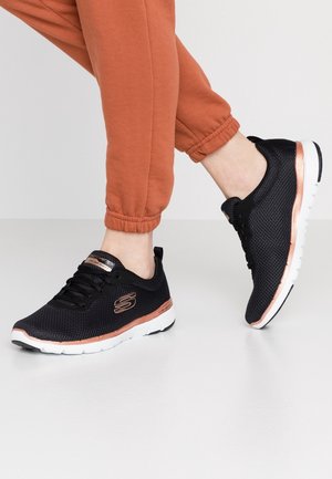 Osoba nosząca czarne siateczkowe buty sportowe z akcentami w kolorze różowego złota i białą podeszwą, w połączeniu z pomarańczowymi spodenkami dresowymi.