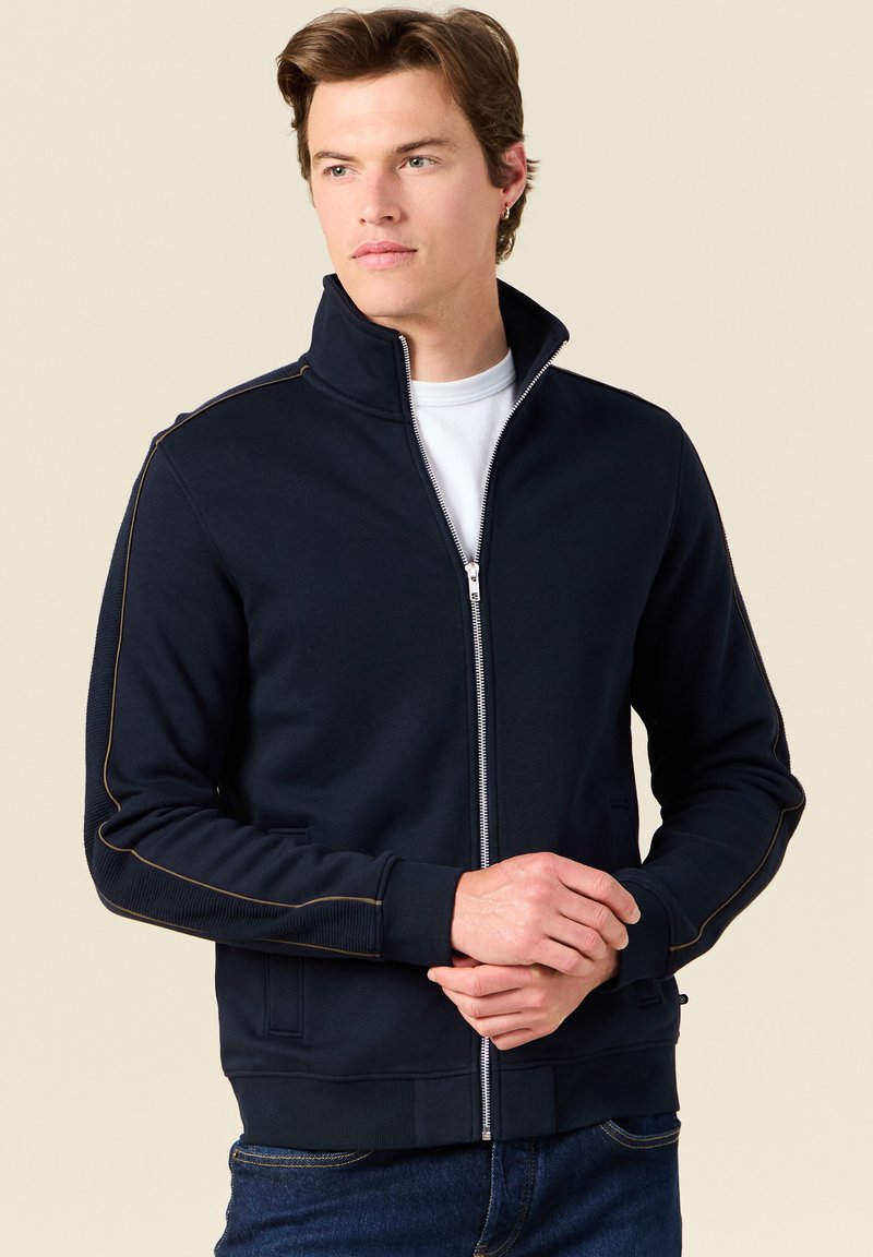 Giacca zip-up blu navy realizzata in tessuto liscio, caratterizzata da un colletto alto, polsini a coste e piping a contrasto lungo le maniche.