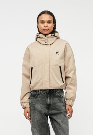 Bežna hladna jakna z kapuco, z žepi na zadrgo, elastičnimi manšetami in majhno logotipno oznako na prsih. Oblečena z temno vzorčastimi kavbojkami.