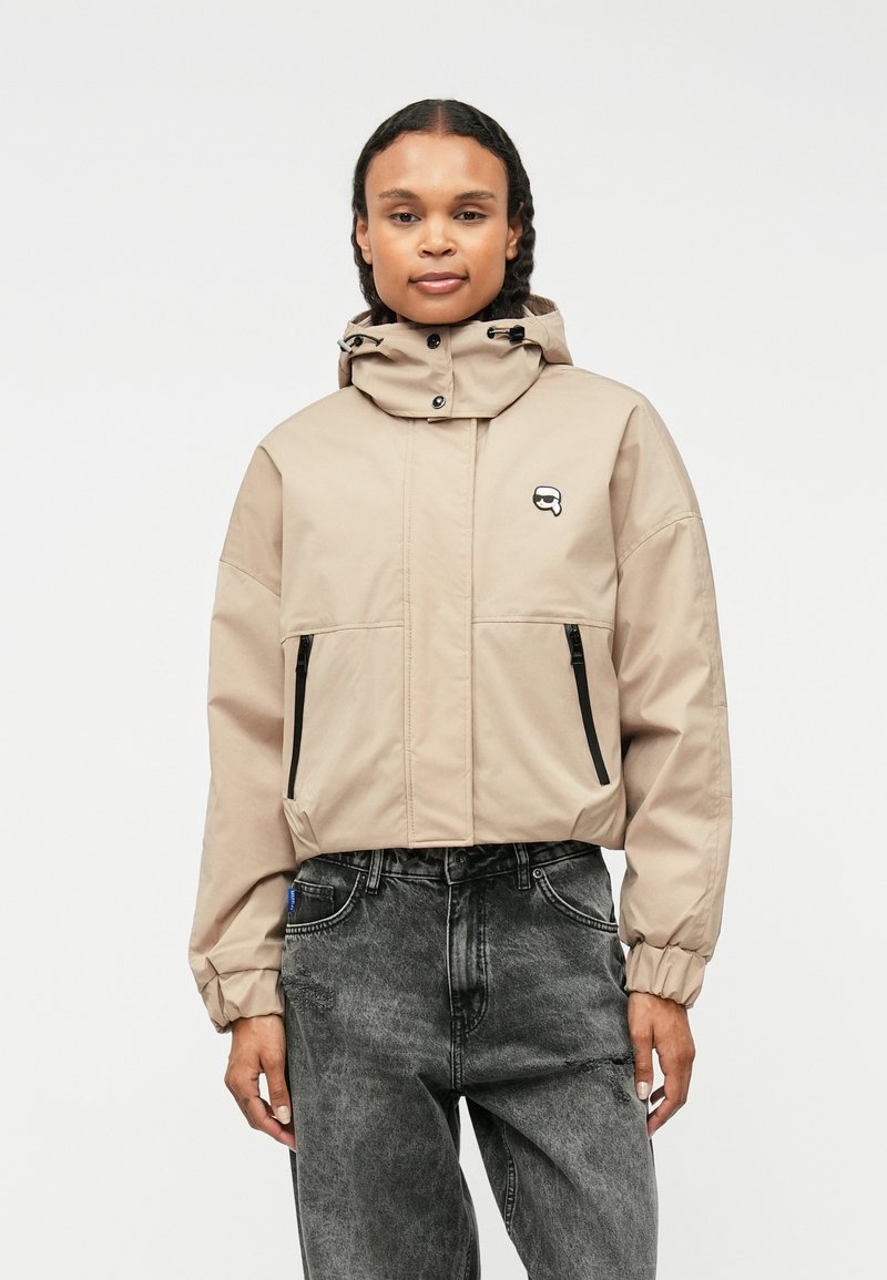 Beige, könnyű dzseki kapucnival, cipzáras zsebekkel, elasztikus mandzsettákkal és egy kis logóval a mellrészen. Sötét mintás farmernadrággal viselve.