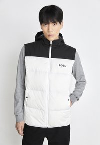 Pūstas liemenė su juodu kapuce ir viršutine dalimi, balta apatine dalimi, pilkomis ilgomis rankovėmis ir priekyje užtrauktuku. Yra logotipas ant krūtinės.