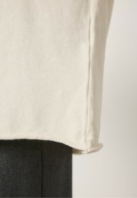 Kremowy sweter o gładkiej fakturze, z zaokrąglonym dołem i widocznymi detalami szwów, prezentowany na ciemnym manekinie.