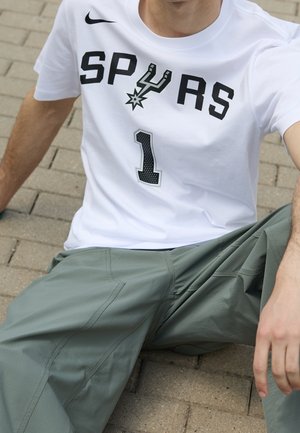 Osoba siedząca na chodniku, ubrana w białą koszulkę Spurs z czarnym napisem i numerem 1, w połączeniu z zielonymi casualowymi spodniami.