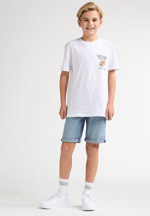 Wit katoenen T-shirt met een grafische print op de borst, gecombineerd met lichtblauwe denim shorts. Model draagt witte sneakers en gestreepte sokken.