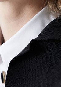 Collar de chaqueta negra superpuesto sobre una camisa blanca. La textura es suave con costuras visibles y un sutil borde deshilachado en el collar.