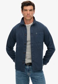 Marineblauwe button-up jas van katoen met een platte kraag, voork pocket en bruine knopen. Draag het over een lichtgrijze trui en denim jeans.