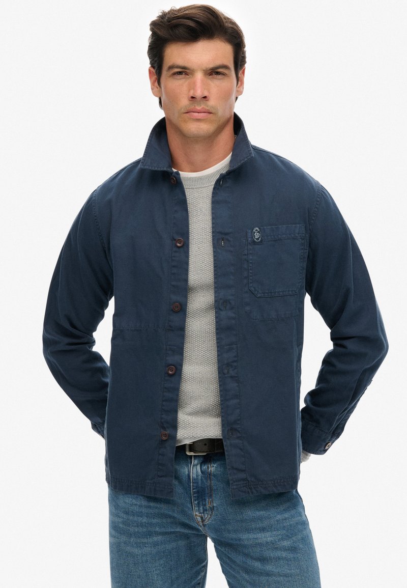 Marineblauwe button-up jas van katoen met een platte kraag, voork pocket en bruine knopen. Draag het over een lichtgrijze trui en denim jeans.