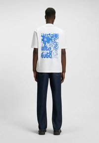 White t-shirt with a blue graphic design featuring text and abstract shapes on the back. The model wears blue jeans and black shoes.
