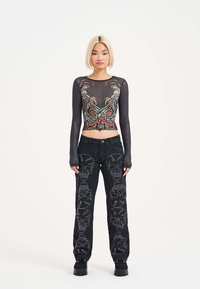 Black sheer long-sleeve top with colorful graphic print, paired with black jeans featuring skull embroidery and a straight-leg design.