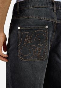 Donker denim jeans met oranje stiksel op de achterzak, die een gestileerd logo-ontwerp heeft. De stof heeft een klassieke textuur.
