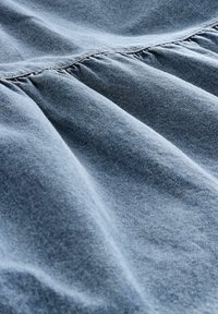 Una tela de denim azul claro con una textura suave, que presenta detalles de costura fruncida. El material tiene un brillo sutil y un tejido irregular.