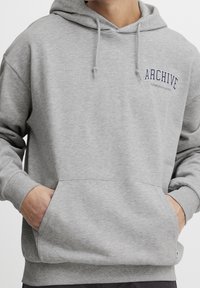 Sudadera gris con bolsillo frontal, capucha ajustable con cordones y logo "ARCHIVE" en color azul marino. Textura suave, corte relajado, puños de canalé.